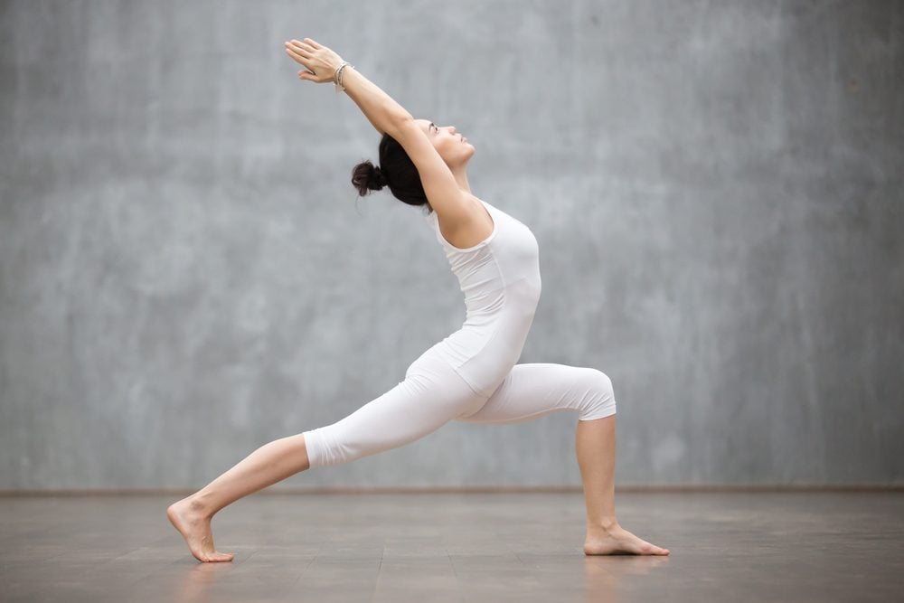 Side view portrait of beautiful young woman wearing white tank top working out against grey wall, doing yoga or pilates exercise. Standing in Warrior one pose, Virabhadrasana. Full length|