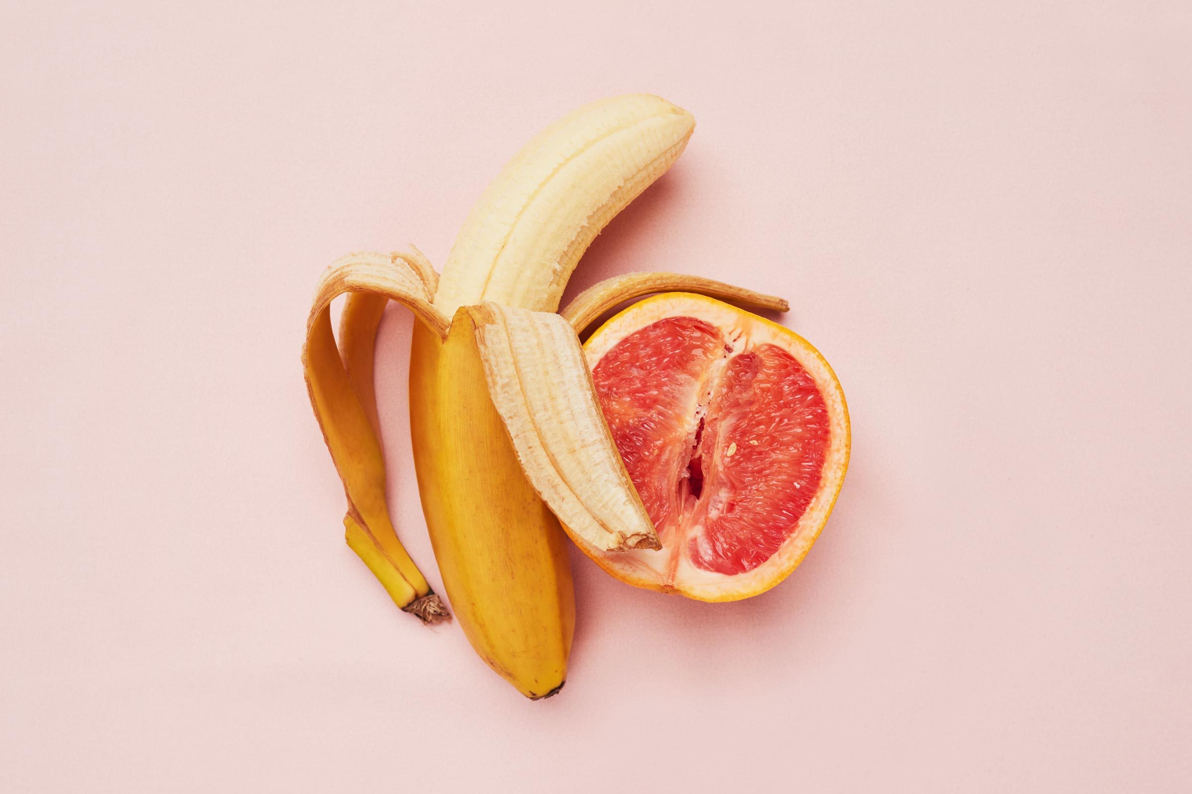 Studio shot of a banana and grapefruit in a suggestive position against a pink background