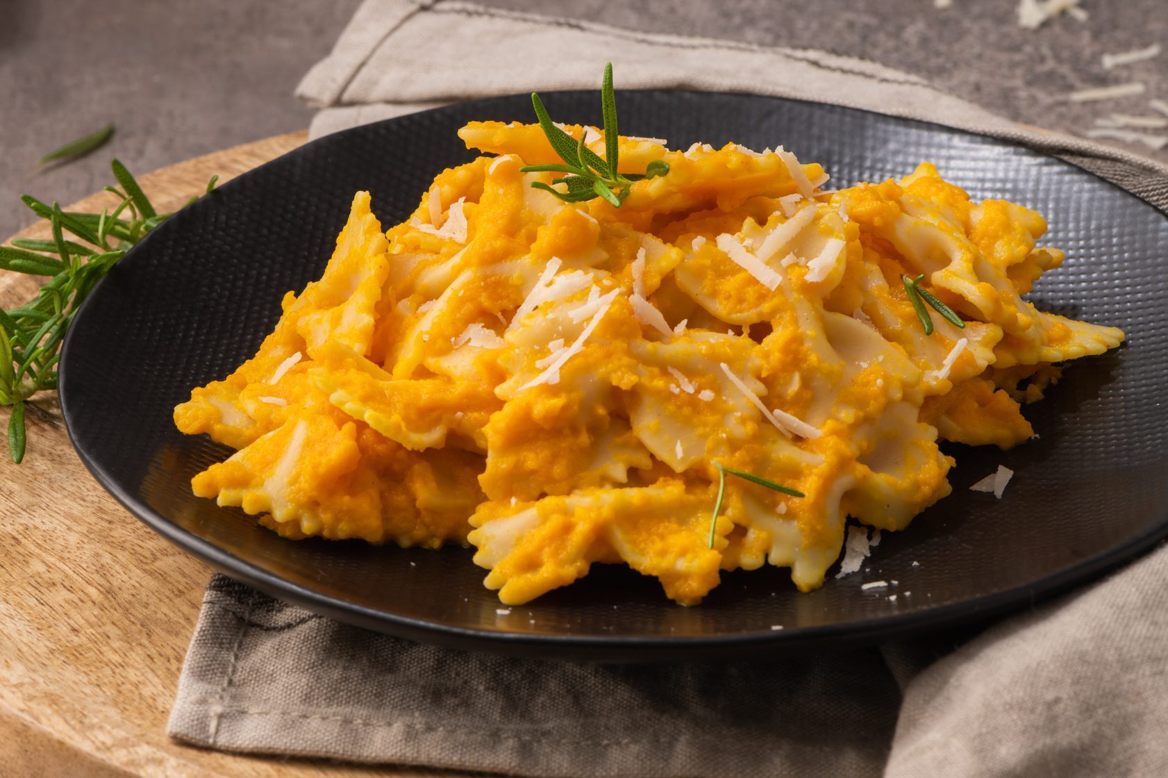 Farfalle pasta with pumpkin sauce and parmesan cheese decorated with rosemary on a black ceramic plate. Creamy cheesy vegan pumpkin pasta. Kitchen counter top.