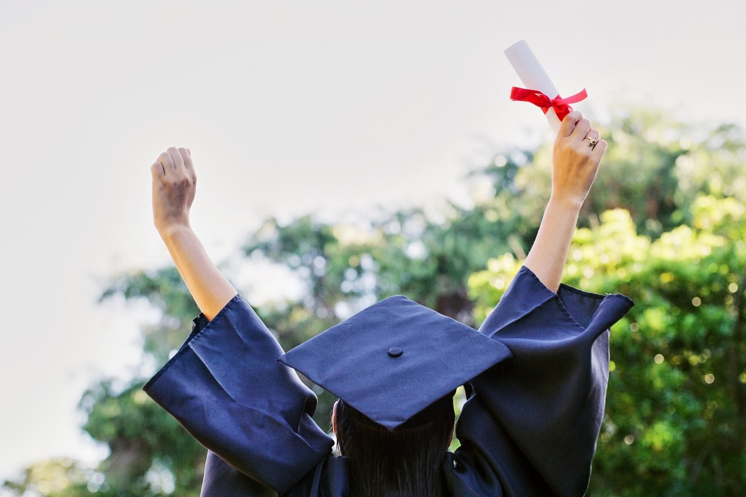woman from behind wearing a cap and gown for graduation and holding up both arms with a rolled up degree in her right hand