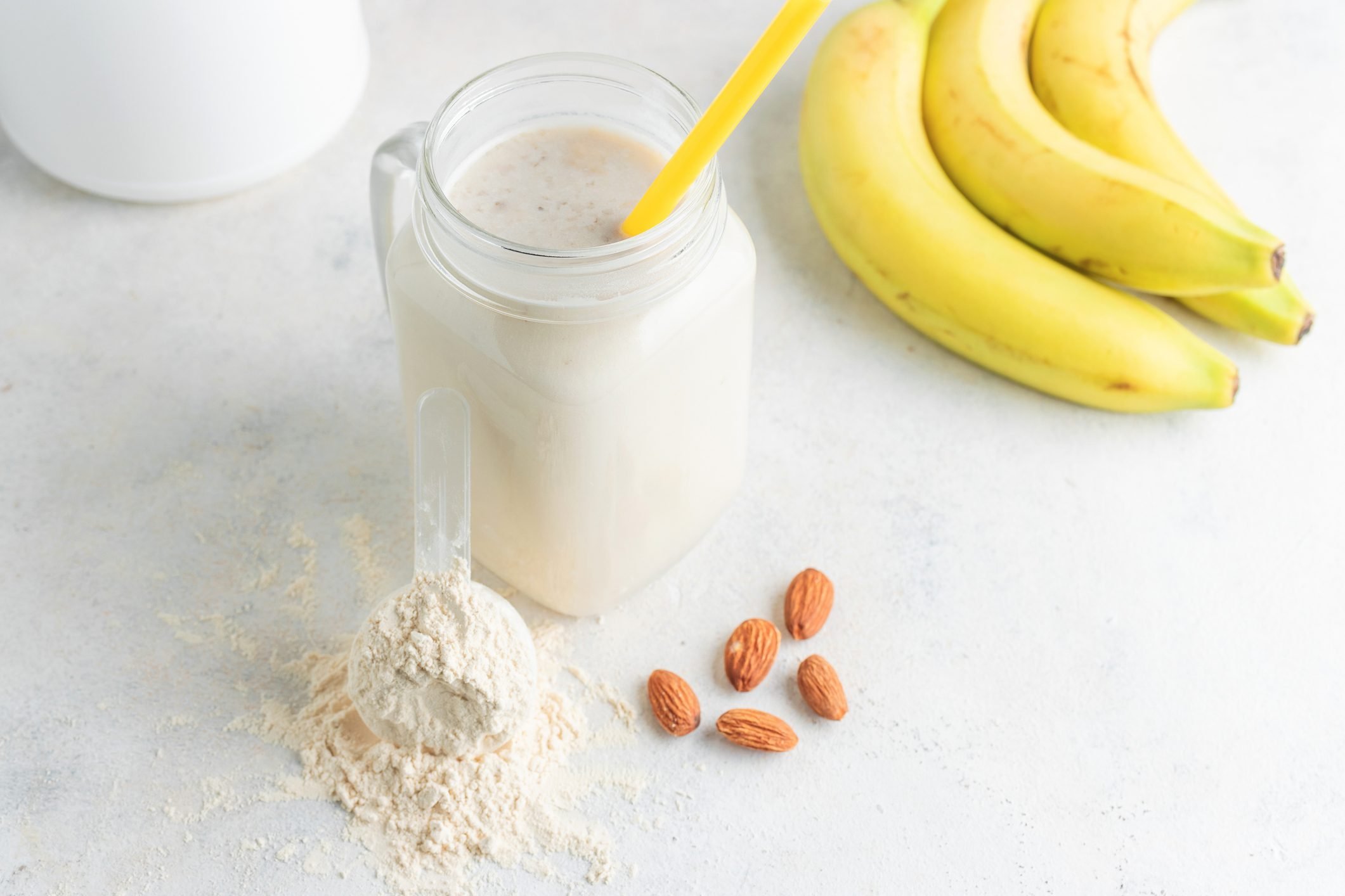 Glass jar of protein milkshake drink protein powder in measuring spoon, bananas, almond nuts on a white background.