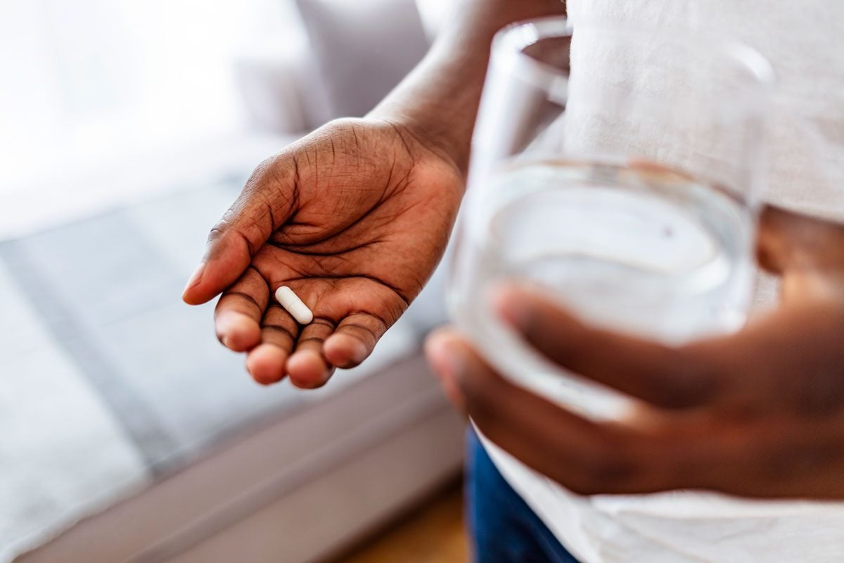 Cropped shot of an unrecognizable person with a pill in their hand, glass of water in the other, about to take a probiotic supplement while standing at home.